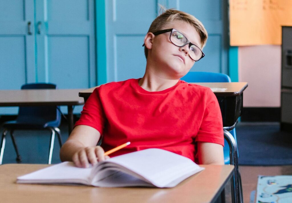A young boy wearing glasses and a red shirt daydreams in an empty classroom, leaning back in his chair.